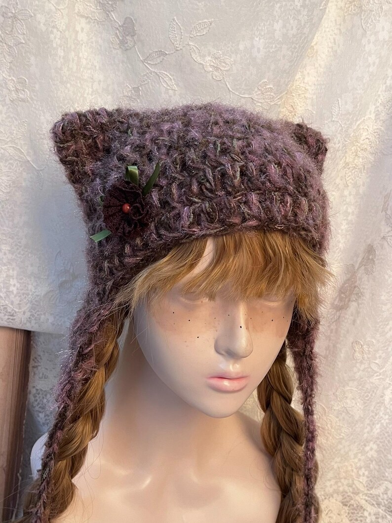 May include: A handmade, purple cat ear hat with ear flaps. The hat is adorned with a dark red flower and green ribbon. The hat is made of a textured, fuzzy yarn. The hat is designed to be worn on the head.