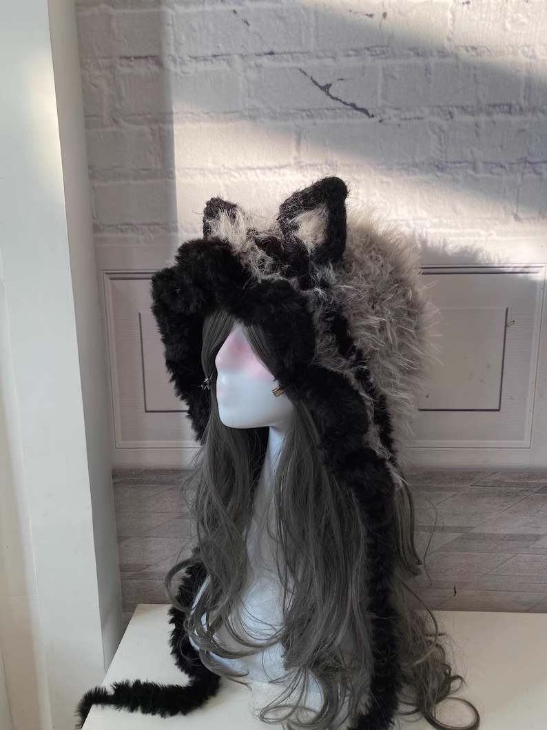 May include: A black and white faux fur hooded hat with long, dangling ear flaps. The hat has a fluffy texture and is designed to resemble a wolf.