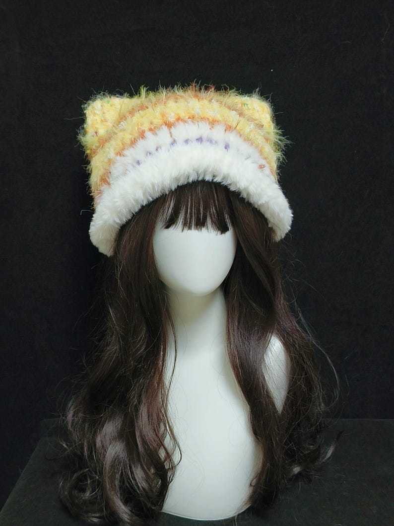 May include: A handmade cat ear hat with a fluffy, textured design. The hat features a mix of yellow, orange, and white yarn, with a white fluffy trim. The hat is displayed on a mannequin head with long, dark brown hair.