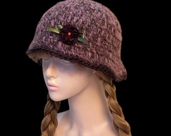 Puede incluir: Un sombrero cloche de punto morado oscuro texturizado. El sombrero presenta una pequeña flor roja oscura con hojas verdes en el lateral. El sombrero tiene una copa redondeada y un ala ligeramente acampanada. El fondo es negro.
