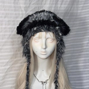 May include: A fuzzy, textured beanie hat in shades of black and grey, featuring long, dangling braided earflaps. The hat is crafted from a mix of soft, shaggy yarn fibres, creating a layered, multi-dimensional appearance with a thick, plush texture.