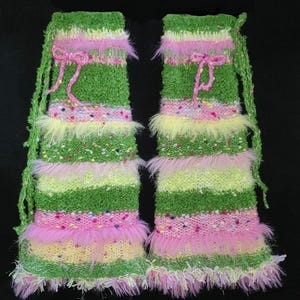 May include: A pair of colorful leg warmers. The leg warmers are green, pink, and yellow with a fuzzy trim. They have a pink bow and a green string. The leg warmers are made of a knitted material.