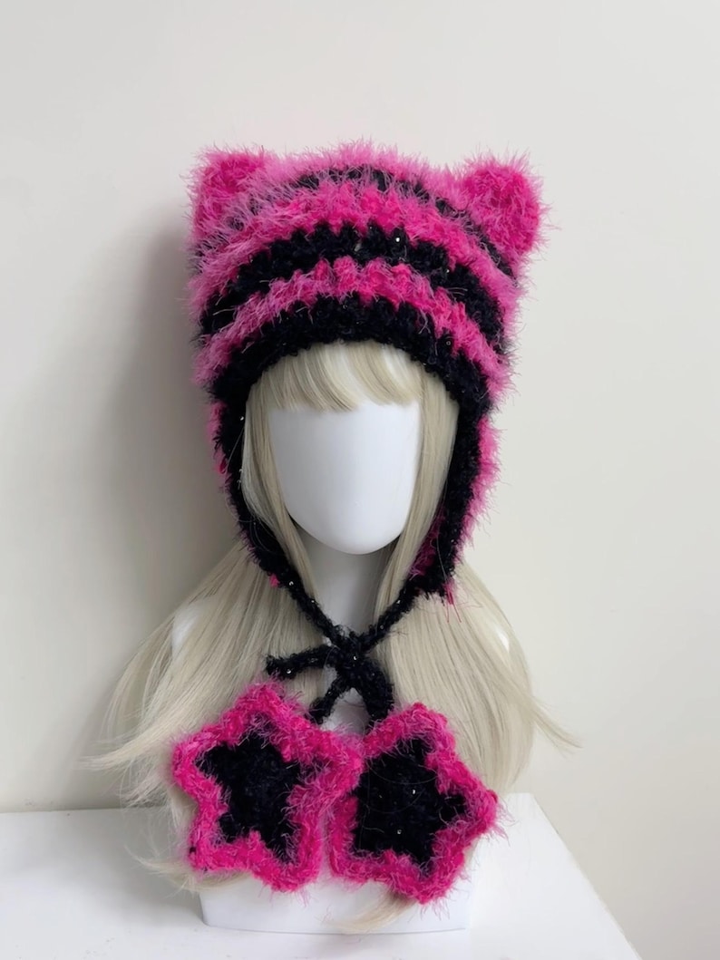 K&ouml;nnte beinhalten: Handgemachtes Set aus einer pink-schwarz gestreiften Katzenohrm&uuml;tze und passenden sternf&ouml;rmigen Fingerhandschuhen. Die M&uuml;tze hat Ohrenklappen und Bindeb&auml;nder unter dem Kinn. Die Handschuhe sind schwarz mit pinkfarbenen Sternkonturen.
