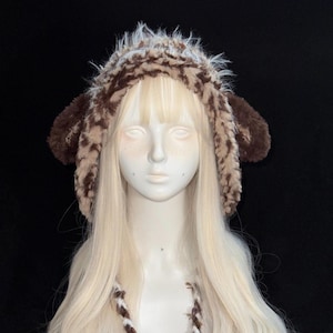 May include: A fuzzy, brown and cream-colored animal-ear hat made of soft, textured faux fur. The hat features rounded ear flaps on both sides and long, braided ties hanging down from the front, designed with a shaggy, multi-toned pattern.