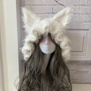May include: A white faux fur hat with large, upright ears, resembling a fox or wolf. The hat is fluffy and covers the head, with long, wavy gray hair cascading down. The hat is displayed on a mannequin head.