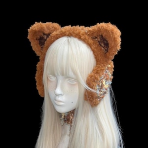 Pode incluir: Uma faixa de cabelo com orelhas de urso marrons e fofas com um colar a condizer. As orelhas têm detalhes em marrom mais escuro. A faixa de cabelo e o colar têm uma aparência texturizada e macia, com tons pastéis.