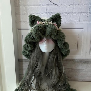 May include: A whimsical, green faux fur cat ear hat with pink floral accents. The hat features large, fluffy ears and long, dangling ties. The hat is displayed on a mannequin head with long, gray hair, against a white brick wall.