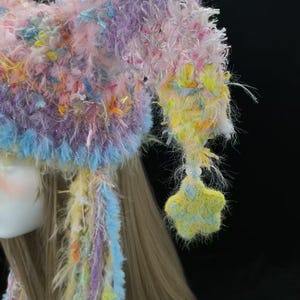 May include: A colourful, handmade hat in pastel shades. The hat has a fluffy, textured design with pink, purple, yellow, and blue hues. A star-shaped decoration hangs from the side. The hat is likely for costume or novelty use.