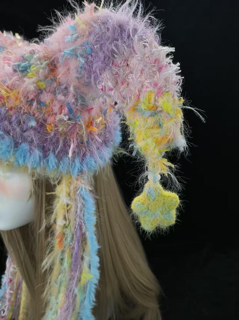 May include: A colourful, handmade hat in pastel shades. The hat has a fluffy, textured design with pink, purple, yellow, and blue hues. A star-shaped decoration hangs from the side. The hat is likely for costume or novelty use.