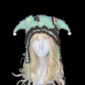 May include: A fuzzy mint green crochet jester hat with dark brown scalloped trim, two pointed side flaps, and dangling star-shaped tassels. The hat features small decorative star and character embellishments and cream-colored pom-poms at the tips.