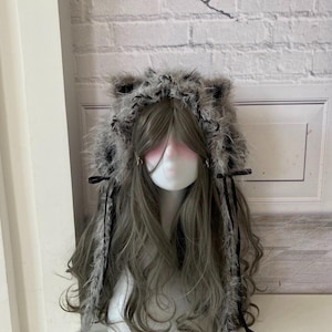 May include: A grey wig with long, wavy hair is styled on a mannequin head. The wig features a furry hood with cat ears in shades of grey and black. Black ribbons and accents are attached to the wig, adding detail to the design.