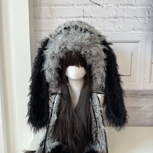 May include: A black and gray faux fur hat with long, floppy ears and a hood. The hat has a fluffy texture and is tied with black ribbons. The hat is displayed on a mannequin head with long, dark hair.