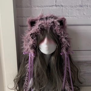 Puede incluir: Un sombrero esponjoso de color lavanda con orejas de gato y pelo largo y gris. El sombrero está adornado con cintas y plumas de color lavanda, creando un accesorio caprichoso y único. El sombrero está diseñado para ser usado.