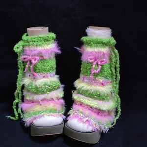 May include: A pair of handmade, colorful leg warmers. The leg warmers are green, pink, and yellow, with a fluffy texture and a pink bow. They are placed over white platform shoes with a brown sole.