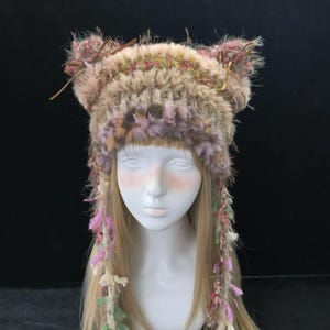 Puede incluir: Un sombrero caprichoso de piel sintética con protuberancias en forma de orejas y hebras colgantes. El sombrero presenta una mezcla de tonos marrón, rosa y verde, con un aspecto texturizado. Las hebras colgantes están adornadas con acentos rosas, verdes y crema. El sombrero se exhibe en una cabeza de maniquí.