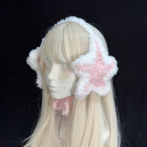 Op de afbeelding: Witte en roze oorwarmers in de vorm van een ster met een bijpassende roze strik. De oorwarmers zijn zacht en pluizig, met een witte hoofdband. Het sterontwerp is roze met witte rand. De strik is bevestigd aan een zilveren ketting.
