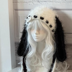 以下が含まれることがあります： 白と黒のふわふわしたニット帽で、長い耳のフラップが付いています。帽子には、前面に小さな黒と白のパンダの顔のディテールがあります。