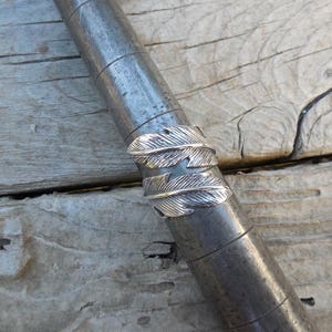 May include: A silver ring with a design of two overlapping feathers. The ring is shown on a metal sizing stick.
