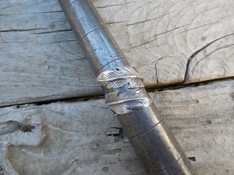 May include: A silver ring with a design of two overlapping feathers. The ring is shown on a metal sizing stick.