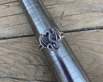 May include: A silver ring with a detailed dragon design. The dragon is facing forward with its wings spread out. The ring is on a metal sizing tool.