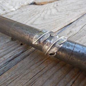 May include: Silver ring with a double feather design. The ring is shown on a wooden surface.