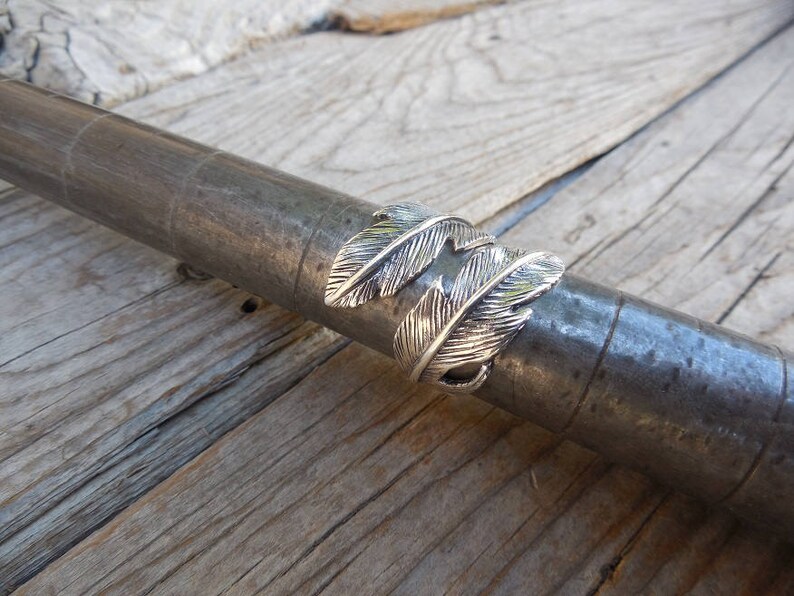 May include: Silver ring with a double feather design. The ring is shown on a wooden surface.