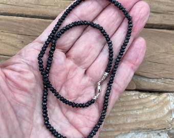 May include: A delicate black beaded necklace with a silver clasp. The necklace is made of small, faceted black beads, creating a shimmering effect. The clasp is a simple, secure design. The necklace is displayed on a wooden surface.