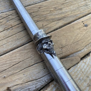 Könnte beinhalten: Ein silberfarbener Metallring mit einem Totenkopf, der einen Zylinder trägt. Der Totenkopf hat detaillierte Gesichtszüge und ist an einer zylindrischen Metallstange befestigt. Der Hintergrund ist eine Holzoberfläche.