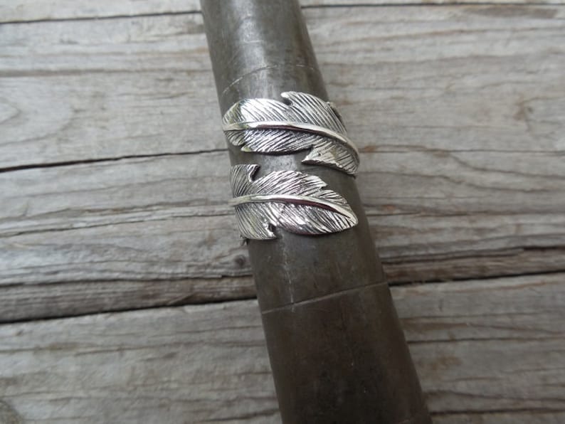May include: A silver ring with two feather designs. The ring is on a gray metal surface.