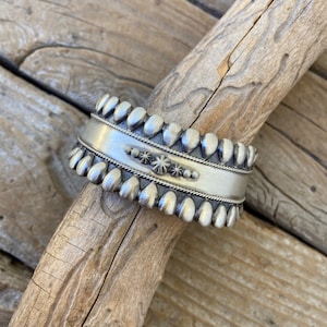 May include: A silver bracelet with a beaded design and a central starburst pattern. The bracelet is on a wooden surface.