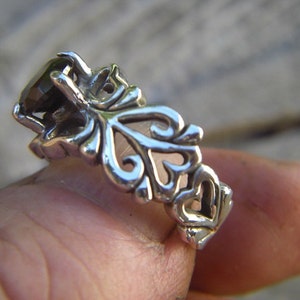 May include: A silver ring featuring a dark gemstone set within an intricate filigree design. The ring is worn on a finger, showcasing the delicate detail of the craftsmanship.