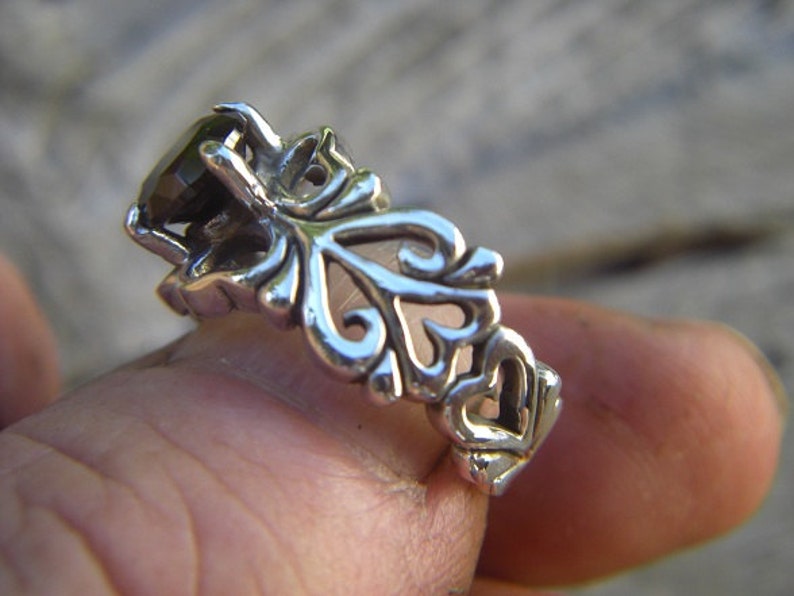 May include: A silver ring featuring a dark gemstone set within an intricate filigree design. The ring is worn on a finger, showcasing the delicate detail of the craftsmanship.