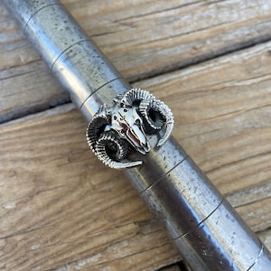 Może przedstawiać: Srebrny pierścionek z detalicznym wzorem czaszki barana. Pierścionek znajduje się na metalowym narzędziu do doboru rozmiaru.