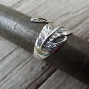 May include: A silver ring with a detailed feather design. The ring is open and adjustable.