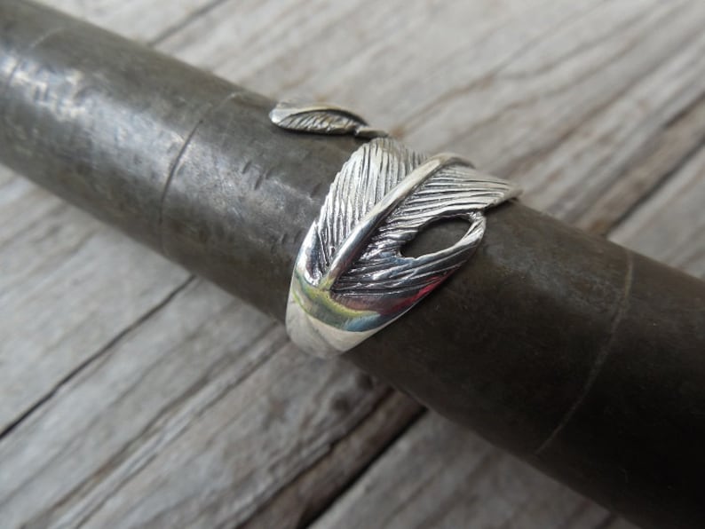 May include: A silver ring with a detailed feather design. The ring is open and adjustable.