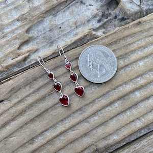 May include: A pair of silver dangle earrings with red heart-shaped charms. The earrings are about the size of a quarter.