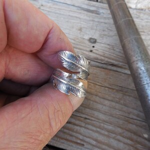 May include: A silver ring with a feather design. The ring is worn on a finger.