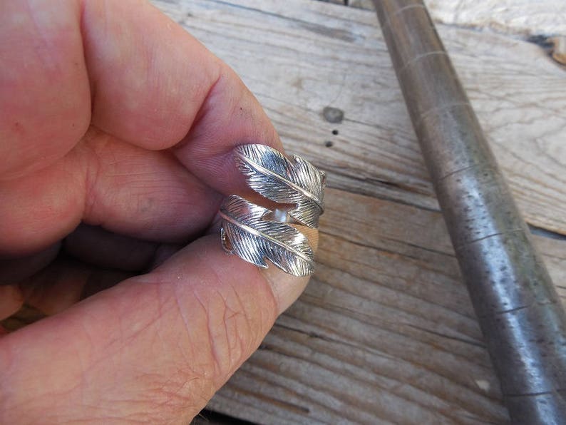 May include: A silver ring with a feather design. The ring is worn on a finger.
