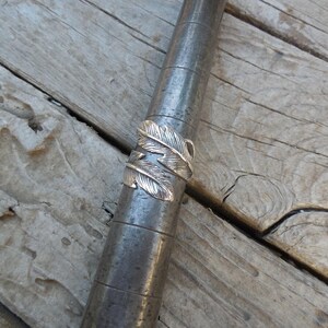 May include: A silver ring with a feather design. The ring is on a wooden surface.