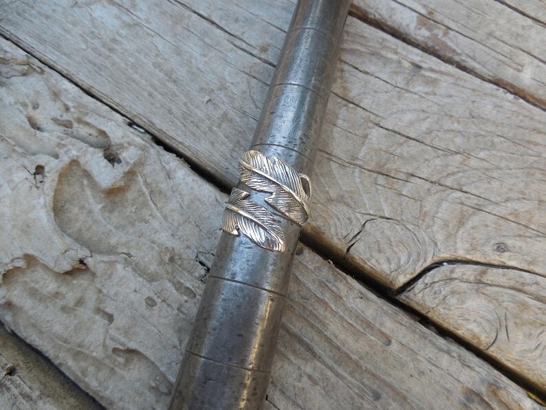 May include: A silver ring with a feather design. The ring is on a wooden surface.
