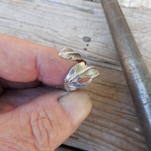 May include: A silver ring with a feather design. The ring is worn on a person's finger.