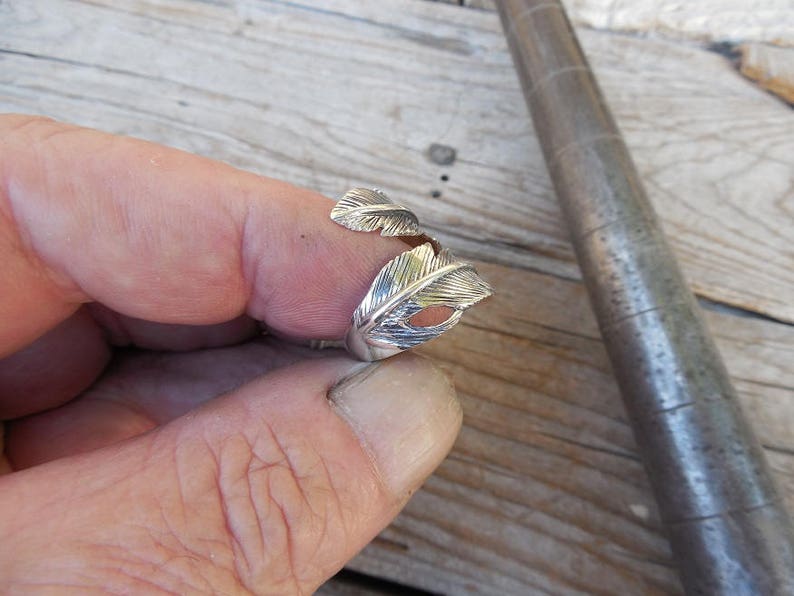 May include: A silver ring with a feather design. The ring is worn on a person's finger.