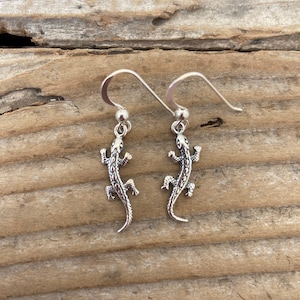 May include: A pair of silver lizard earrings with a dangling design. The earrings are on a wooden surface.