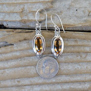 May include: A pair of silver dangle earrings with oval-shaped topaz gemstones. The earrings are about the size of a US dime.