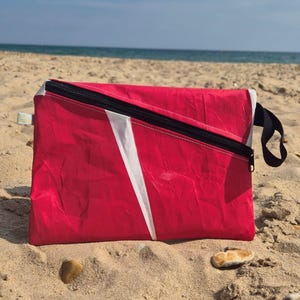May include: A rectangular zippered pouch in bright pink with a diagonal white stripe across the front. The pouch features a black zipper closure running along the top edge and a black fabric loop handle attached to the right side of the bag.