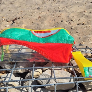 May include: A clear pencil case with a red, green, and yellow fabric top. The case has a yellow zip and a yellow fabric handle. The case is on a metal grid with rocks and sand in the background.