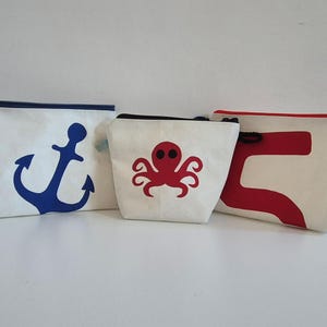 Könnte beinhalten: Drei weiße Reißverschlusstaschen in verschiedenen Größen. Die linke Tasche zeigt ein großes blaues Anker-Motiv. Die mittlere Tasche ist mit einem roten Kraken-Design versehen. Die rechte Tasche zeigt eine große rote abstrakte Form.