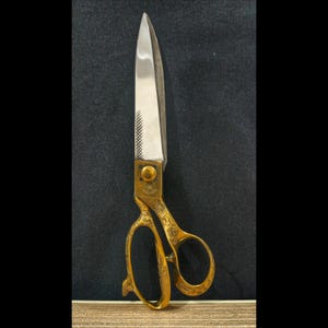 Puede incluir: Tijeras de sastre vintage con una hoja de acero pulido y mangos dorados ornamentados. Las tijeras tienen una punta afilada y un patrón decorativo en los mangos. Las hojas miden aproximadamente 25 cm de largo.