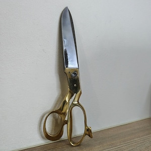 Pode incluir: Tesoura de alfaiate grande, de estilo vintage. As lâminas são prateadas e as alças douradas. A tesoura está encostada numa parede branca e numa superfície de madeira. Mede aproximadamente 25 cm de comprimento.