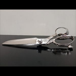 Pode incluir: Um par de tesouras prateadas ornamentadas com uma lâmina polida. As alças apresentam intrincados desenhos florais e orifícios arredondados para os dedos. As tesouras são projetadas para cortar tecido ou papel. Comprimento: aproximadamente 25 cm.